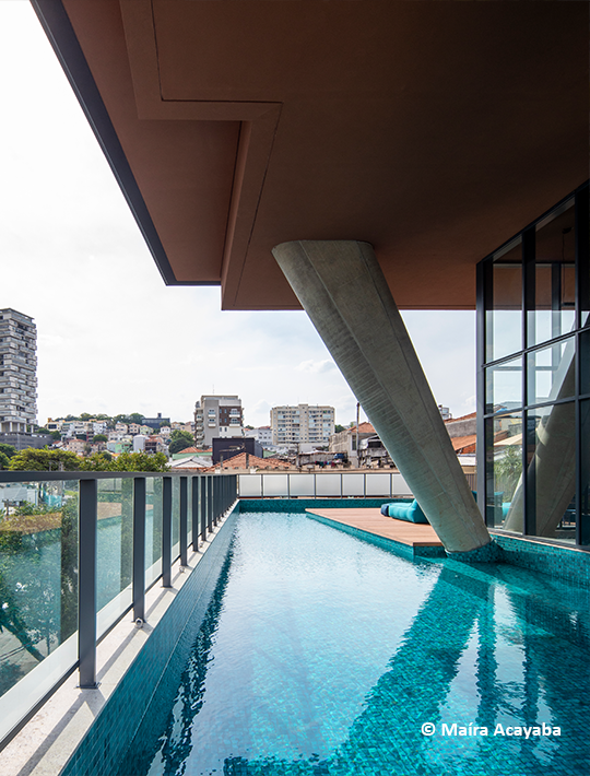 Foto da sala de leitura do edifício Float. No primeiro plano, há dois sofás brancos. No segundo plano, há uma estante de livros. Atrás, a parede de vidro mostra a vegetação do fundo.